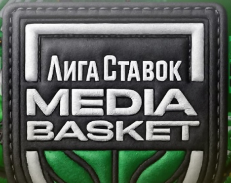 На изображении логотип «Лига Ставок Media Basket». Черно-белый знак на зеленом фоне, состоит из текста и графического элемента, напоминающего баскетбольное кольцо