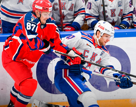 На изображении хоккейный матч: игроки в красной и бело-синей форме борются за шайбу у борта. На заднем плане видны запасные игроки в форме с логотипом СКА. Активная игра, напряженная борьба